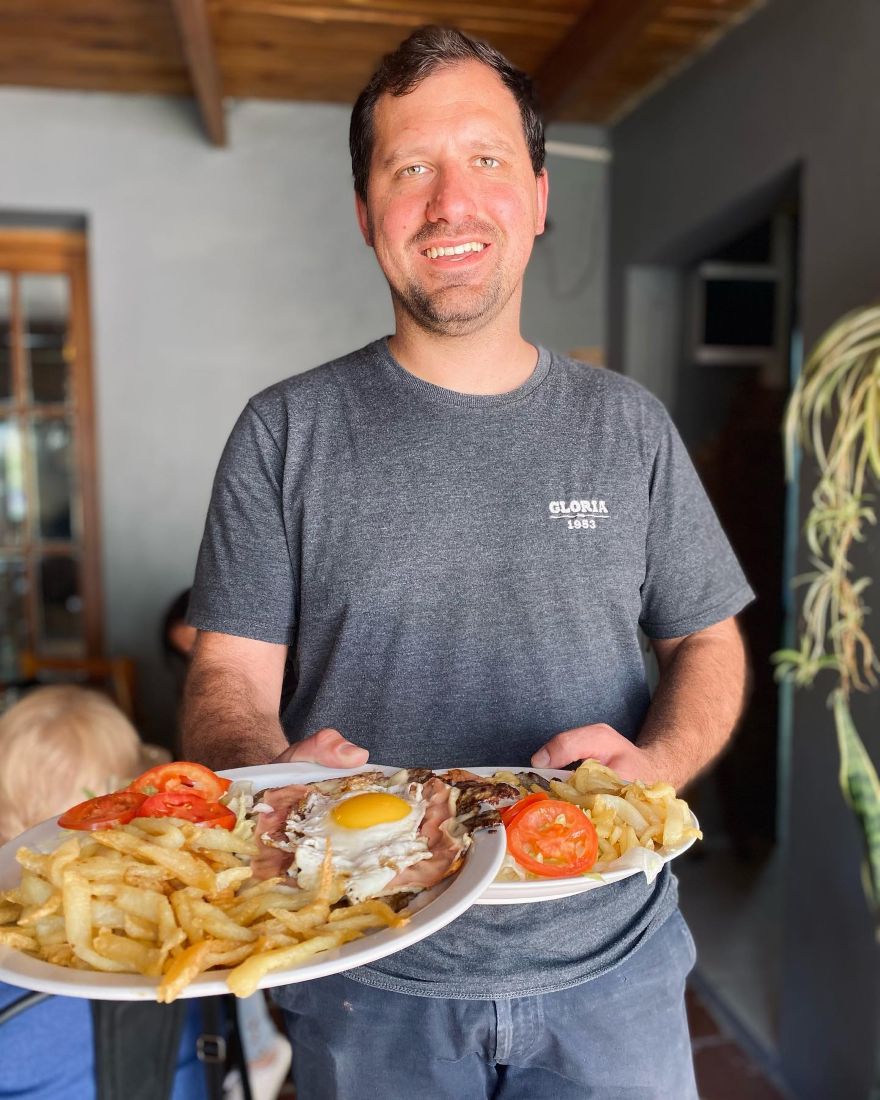 Mozo sonriendo con dos platos de bife con huevo frito, tomate y papas fritas